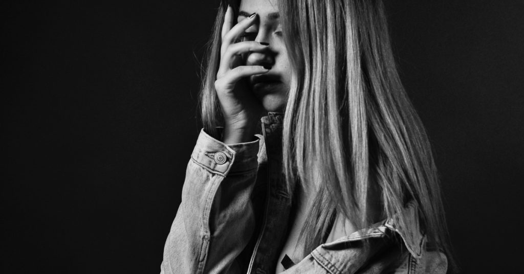 A black and white image shows a young woman clutching her face, in a moment of self-loathing.