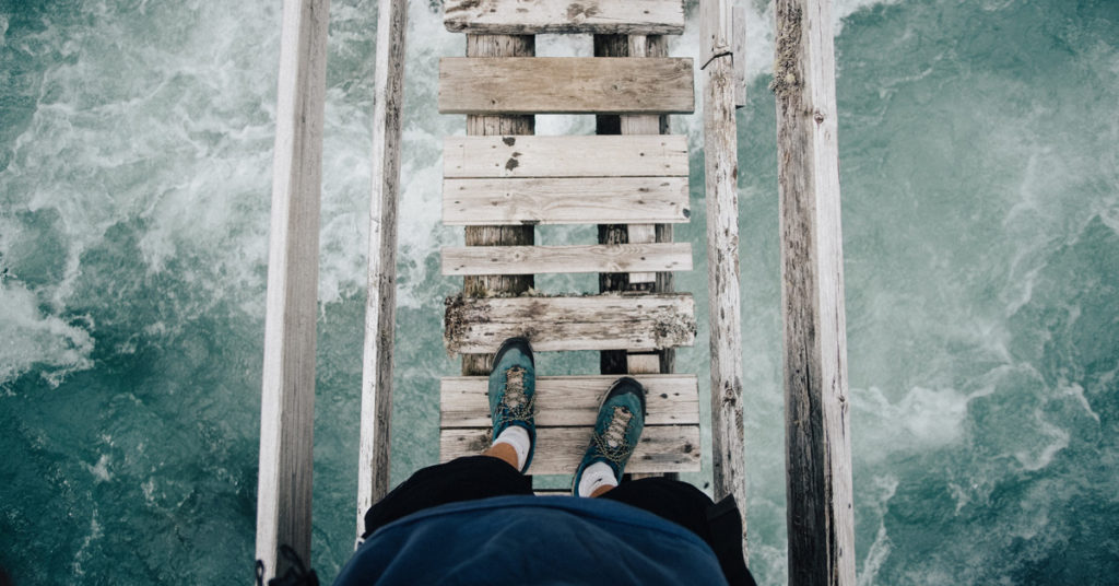 A man in shorts and green shoes stands on a rickety wooden bridge over a rough ocean, showing the difficulty of facing feelings.