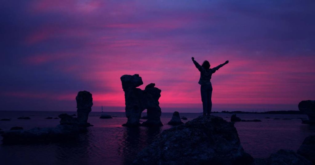 a woman stands on a rock at sunrise, stretching out her arms to show her ambition.